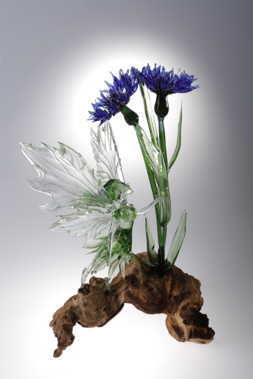 A flameworked glass sculpture featuring a fairy with a cornflower mounted on wood.