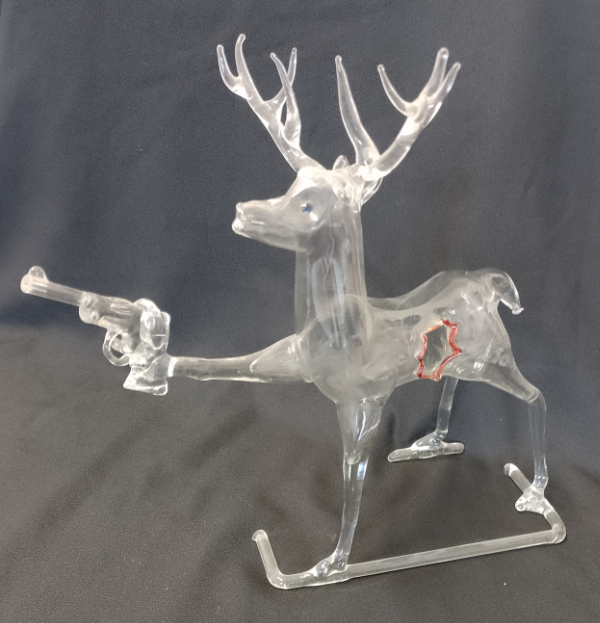 Humorous lampworked clear glass stag with a hole in its side outlined in red, holding a revolver pointing forward in its front hoof. Titled Stag Hunting.