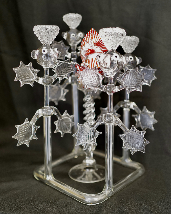 Lampworked clear glass sculpture titled 'Thistle Family at Teacake Barbecue' with four thistle figures standing round a central glass podium with a teacake wrapper on the top. 