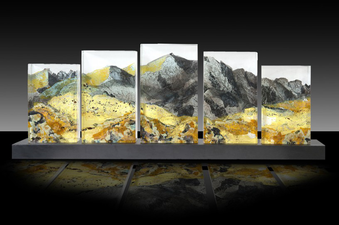 A series of 5 vertically rectangular flat glass panels, with the tallest in the centre and descending sizes of panel each side, mounted on a dark grey base. Painted across the 5 panels is a scene of mountain peaks in tones of grey, yellow and ochre, representing autumn in the Snowdonian Massif.
