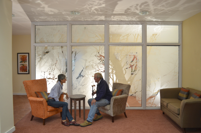 Two people sitting chatting in front of indoor 'Cysgod y Coed' window, Bro Cwm Cerwyn, Crymych, Pembrokeshire, made by Studio Melyn, 2017.