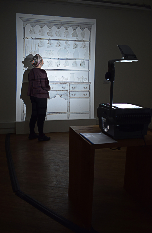 Artist Linda Norris standing with her installation piece Fragment Dresser in a darkened room with light projection on the dresser where clear glass etched cups, jug and teapot are displayed.