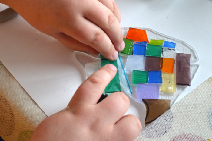 Close up of a child's hands placing coloured glass pieces on a fish-shaped clear glass base. The child is autistic and is being assisted by Linda Norris.