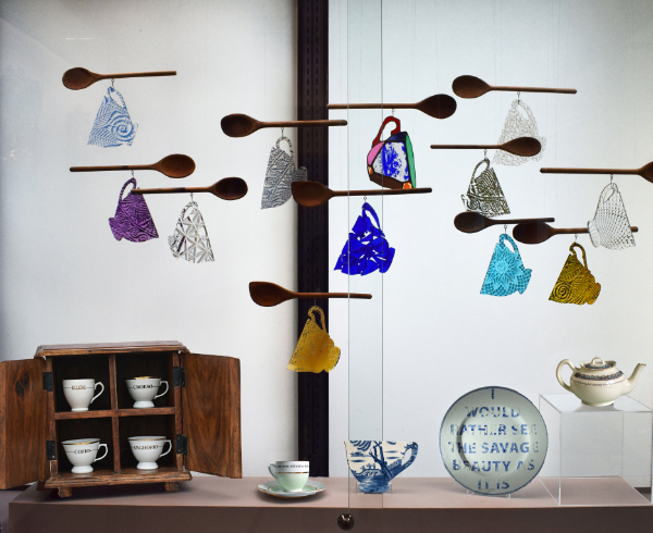 Re:Made exhibition featuring wooden spoons and flat glass tea cups with text on them suspended over a table with other cups, a teapot and plate.