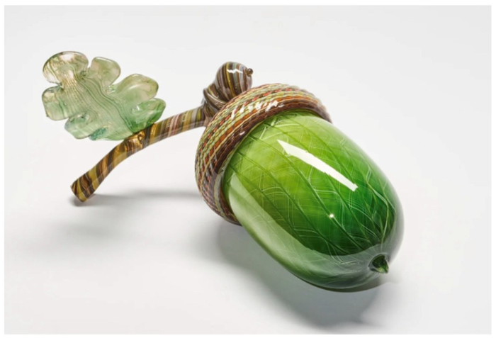 A large green acorn made of glass with a short stem and acorn leaf attached. There are delicate swirled patterns running from the tip of the acorn towards the brown acorn cup and the green tone becomes lighter towards the cup. 