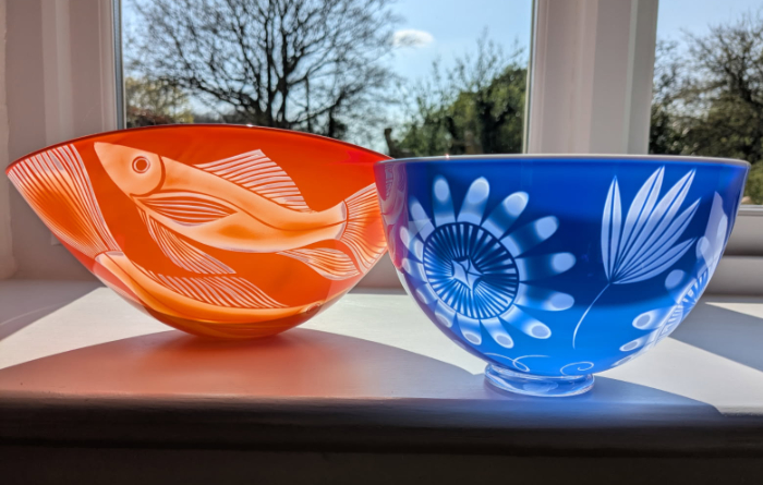 Two sandblasted glass bowls, one in orange featuring swimming fish and the other in dark blue featuring a flower and leaf design.