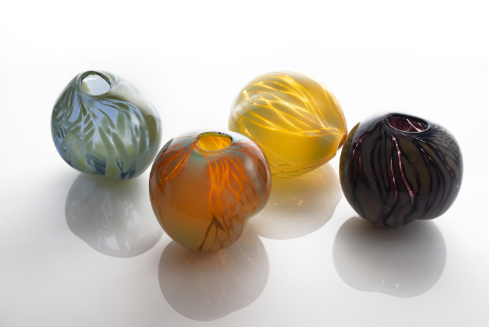 Four uneven shaped, rounded glass pods in different colours with etched designs cut into the surface to reveal clear glass below.