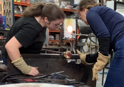 Glass artists Maria Zulueta and Lucy Richard working together on molten glass blob on a pipe.