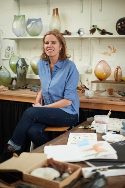 Michele Oberdieck seated in her glass studio surrounded by glass vessels and objects and with painted designs in sketchbooks in the foreground.