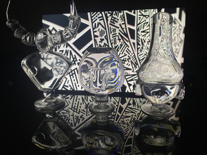 Clear glass masks made by Theo Brooks standing in front of light box projecting geometric patterns which are reflected and refracted through the masks.