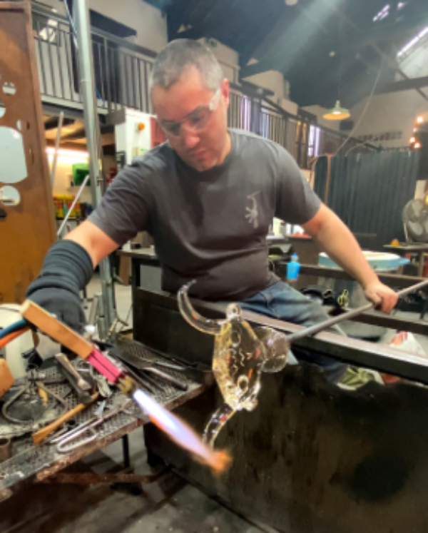 Theo Brooks sitting in the glass hot shop turning a glass mask on a metal pole and heating a part of it with a torch.